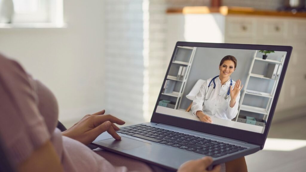 Uma pessoa usa um laptop para uma videochamada com um profissional médico de jaleco branco e estetoscópio, representando a privacidade em consultas online