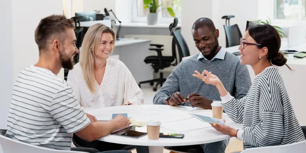 comunicação assertiva Quatro pessoas sentam-se ao redor de uma mesa em um escritório iluminado, envolvidas em comunicação assertiva. Elas seguram papéis e dispositivos eletrônicos enquanto tomam café. A sala tem móveis modernos e janelas grandes, criando um ambiente ideal para discussões eficazes no local de trabalho.