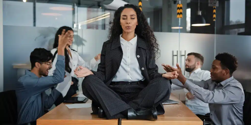 Colaboradores em uma mesa no ambiente de trabalho tendo discussões, com estresse e sobrecarga, mas uma colaboradora está sentada em cima da mesa em posição de lotus pela yoga, fazendo uma meditação e tentando se acalmar. Impacto da NR-01 nas empresas são significativos e de extrema importância para produtividade, saúde e bem-estar do time