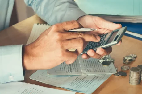 Uma pessoa calcula meticulosamente com uma caneta na mão, cercada por documentos financeiros e pilhas de moedas em uma mesa de madeira, incorporando eficiência tributária com precisão. Perto, um jarro de vidro transbordando de moedas reflete sua dedicação à gestão inteligente de recursos.