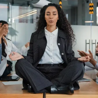 Colaboradores em uma mesa no ambiente de trabalho tendo discussões, com estresse e sobrecarga, mas uma colaboradora está sentada em cima da mesa em posição de lotus pela yoga, fazendo uma meditação e tentando se acalmar. Impacto da NR-01 nas empresas são significativos e de extrema importância para produtividade, saúde e bem-estar do time