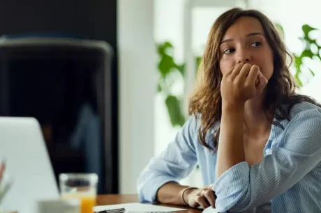 mulher no trabalho preocupada, demonstrando estar cm crise de ansiedade no trabalho. Esta sentada em uma mesa, em frente ao computador com papeis em cima da mesa e olhando para frente com a mão no queixo, demonstrando preocupação e ansiedade