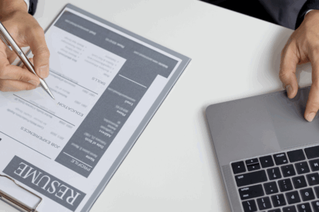 pessoa escreve em um currículo, enquanto sua outra mão paira sobre o teclado do laptop, representando a transição de carreira